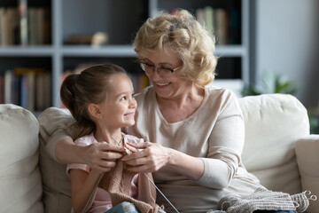 Smiling loving mature grandmother wearing glasses teaching little granddaughter knitting, hugging,...