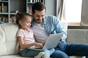 Smiling little girl with father using laptop together at home, sitting on cozy couch in living room, looking at screen, watching video or cartoons, shopping online, chatting, enjoying leisure time