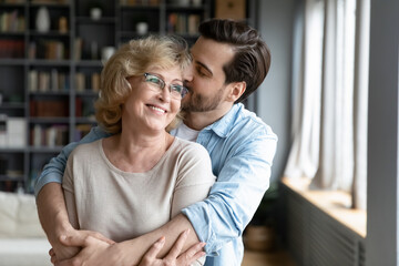 Loving adult son kissing and hugging happy mature mother from back close up, expressing gratitude, middle aged woman wearing glasses and young man enjoying tender moment, standing in living room