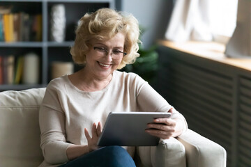 Smiling mature woman wearing glasses using computer tablet at home, positive overjoyed middle aged female browsing apps, shopping or chatting online, sitting relaxing on cozy couch at home