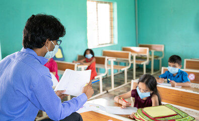 Fototapeta premium Teacher in medical mask with limited kids teaching at classroom while maintaining social distance due to coronavirus or covid-19 pandemic