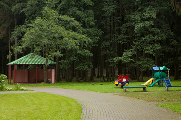 An empty little playground, a gazebo in a garden or park with a green mowed lawn and tall trees.