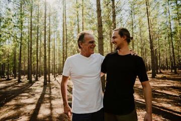 Father and son hugging while smiling at each other standing in green forest.
