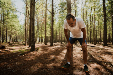 Fit elderly male tired bending over with hands on knees in green forest. 
