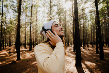 Handsome caucasian male wearing beanie and headphones smiling while listening to music standing in green forest.