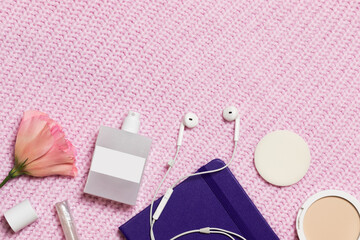 Closed purple notebook on a pink knitted background with copyspace. There are a pink flower, white perfume, white headphones, powder, lip gloss.