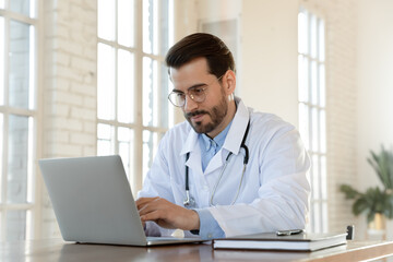 Focused young doctor wearing glasses working on laptop in office, sitting at desk, looking at screen, serious therapist gp writing report or email, consulting patient online, filling medical card