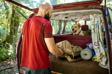 Young bearded man with his dog, labrador retriever, unpacking minivan for camping and hiking in the nature 
