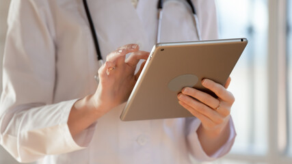 Close up young woman doctor holding computer tablet, using medical apps, chatting, consulting patient online, telemedicine concept, therapist gp wearing white uniform standing in office