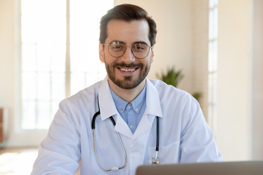 Head shot portrait smiling confident young doctor wearing glasses and medical uniform with stethoscope looking at camera, therapist physician gp sitting at desk with laptop in office