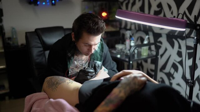 
tattoo artist makes a tattoo on the legs of a young woman who lies on the couch and relaxes