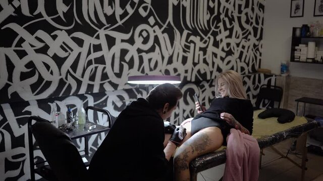 
young woman laughs and uses a mobile phone while getting a tattoo by a professional tattoo artist