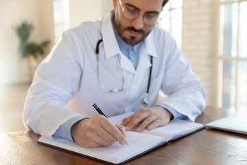 Close up focused doctor wearing uniform working with documents, serious young man therapist physician writing in journal, book, illness history or anamnesis, sitting at work table in office