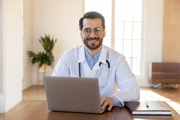 Head shot portrait smiling doctor wearing glasses working on laptop, sitting at desk in office, therapist physician gp wearing white medical uniform with stethoscope looking at camera