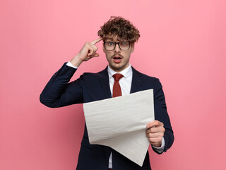 shocked young man in suit pointing finger to temple and reading newspaper
