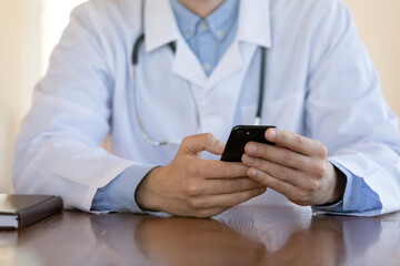 Close up doctor wearing white medical uniform with stethoscope holding phone, browsing medical apps, sitting at work desk, therapist physician gp using smartphone, healthcare and technology concept