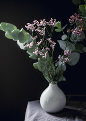 White ceramic vase with eucalyptus brand and violet flowers against a black wall.