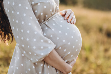 A pregnant woman in a dress holds her hands on her stomach on a nature background. Pregnancy, motherhood, preparation and expectation concept. Close-up, Beautiful gentle mood photo  - 376189759