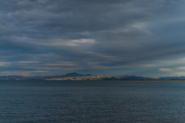 view of the clear calm undulating blue water of Lake Baikal, sunset mountains on the horizon, clouds