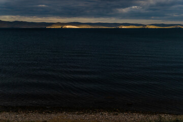 Fototapeta premium view of the clear calm undulating blue water of Lake Baikal, sunset mountains on the horizon, clouds, coast
