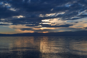 Fototapeta na wymiar view of the clear calm undulating blue water of Lake Baikal, mountains on the horizon, sunset sky, clouds, reflection