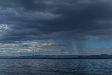 Naklejka na ściany i meble view of the clear calm undulating blue water of Lake Baikal, mountains on the horizon, rain clouds, shower
