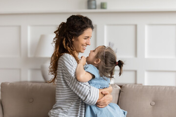 Side view caress mom cuddle little daughter small princess wear crown sit on sofa looking at each other enjoy moment of sincere and unconditional love. Mother Day celebration, happy motherhood concept