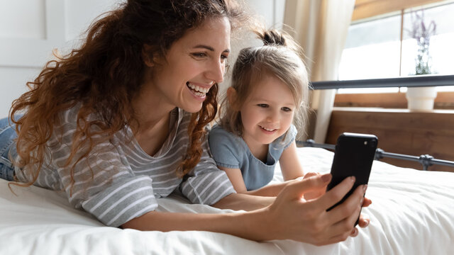 Close up nanny show cartoons on smartphone to little kid girl. Waking up lying on bed mom and daughter use phone make videocall enjoy distant talk with relatives, parental control modern tech concept