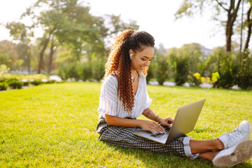 Portrait of pretty young woman sitting on green grass in park with  laptop. Happy woman, student, freelancer with wireless headphones calling on laptop, talk by webcam, video conference.