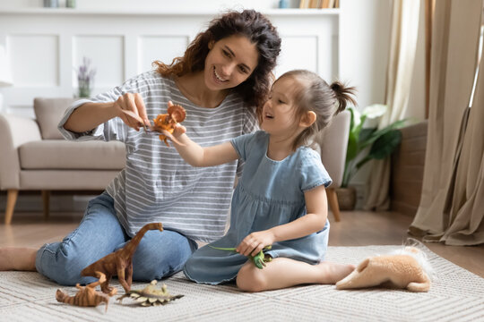 Mom and daughter play with dinosaurs set. Baby sitter and kid girl spend time at home sit on carpet in cozy living room. Enjoy educational game, have fun together, parent develop child, hobby concept