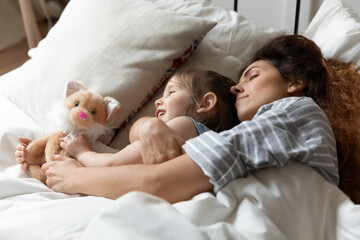 Obraz na płótnie Canvas Mom hug small daughter during daytime nap in bed, close up. Serene kid girl rest lying with cat stuffed animal toy and loving mother with eyes closed under duvet. Family healthy sleep together concept