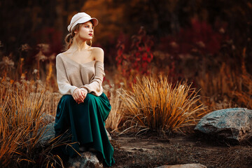 beautiful girl in a cap and coat with a bare shoulder on the background of autumn walk green skirt, sitting