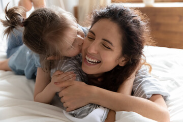 Fototapeta premium Close up face happy mother enjoy time with small daughter in morning in bedroom. On bed cheery kid lively mom laugh play together, child kiss mum cheek, lying on her back, relish tender moment concept