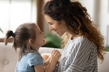 Side view young beautiful mother and little pretty daughter sit on couch at home smile looking at each other with tender and love. Happy motherhood, protection and care. Child or Mother Day concept