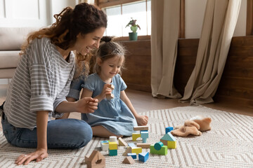 Mom and little daughter play with color wooden bricks toys set. Baby sitter and small kid girl sit on warm floor on carpet enjoy funny game together. Educational activity, development of child concept - obrazy, fototapety, plakaty