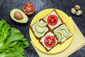 Avocado and tomato sandwich on a yellow plate. Lettuce leaf and quail egg on a dark board. Vegetarian food, dieting nutrition concept, still life. Toasted bread for lunch. Flat composition, top view.
