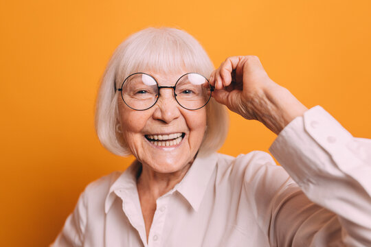 ?lose-up senior woman with blonde short hair wearing light blouse, smiling and fixing her glasses by her hand. Woman isolated over bright orange background.