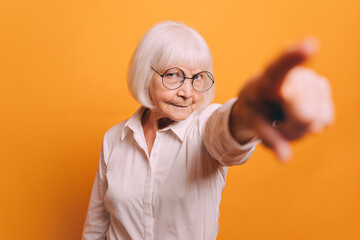 Dissatisfied senior woman with blonde hair wearing round glasses and light blouse standing isolated over orange background and pointing a blurred finger at something behind camera.