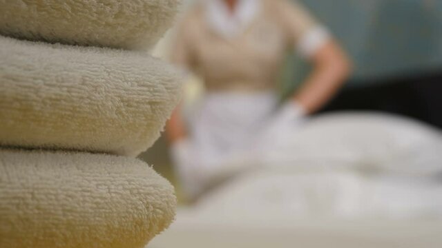 Hotel concept. Blurred shot of maid in uniform and apron making bed in room, shot through stack of fresh bath towels on bed sheet. Preparation of hotel room. 4 k video