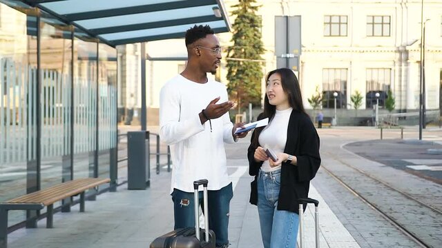 Handsome smiling bearded african amercian talking with his joyful asian female friend while they waiting on public transport on tram station with suitcases