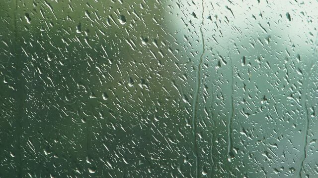 Rain drops sliding slow on window glass in rainy day, de-focused green tree in background, medium closeup shot