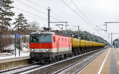 Fototapeta na wymiar A large, powerful electric locomotive in red and white is pulling a train of freight cars. Passenger platform. Winter snowy weather.
