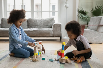 Happy african american older sister involved in constructing buildings with younger little biracial brother in living room. Smiling adorable mixed race small children siblings playing toys at home.
