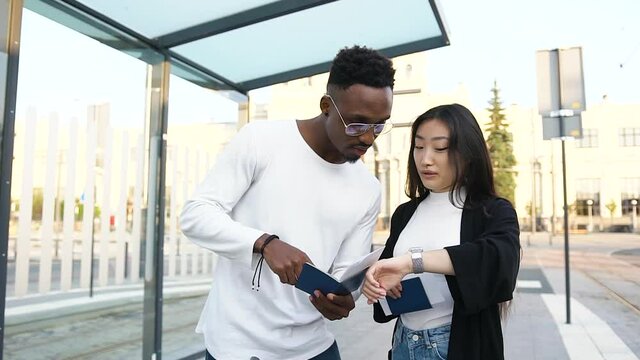 Attractive positive young multiracial friends checking travelling documents,then controlling time and carrying their suitcases on public tram station near railway