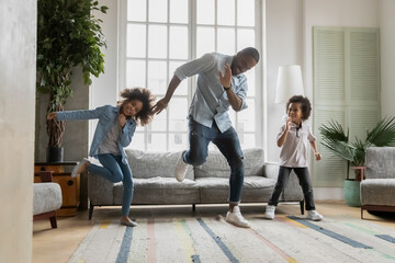 Happy african american father showing funny dancing moves to energetic small children siblings at home. Laughing crazy little biracial boy and girl having fun with caring daddy in living room. - Powered by Adobe