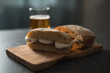 Ciabatta sandwiches with salami and mozzarella on olive wood board next to glass of beer