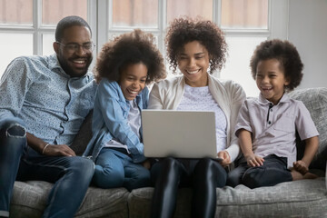 Smiling young biracial woman holding on laps computer, watching funny movie or comedian cartoons with joyful small older daughter, younger son and handsome husband, happy family weekend pastime. - Powered by Adobe