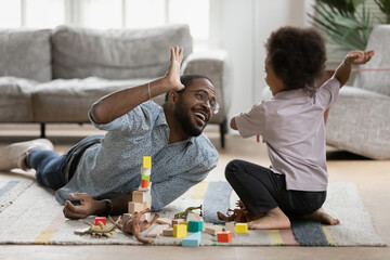 Happy young handsome biracial father lying on floor carpet, giving high five to joyful little...