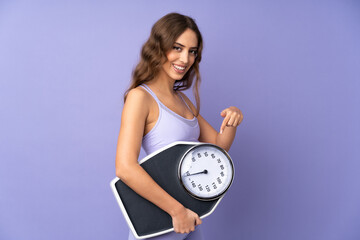 Young sport woman over isolated purple background holding a weighing machine and pointing it