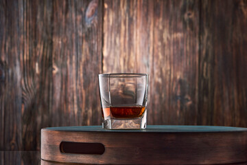 A square glass of whiskey stands on a tray against an old wooden wall.
